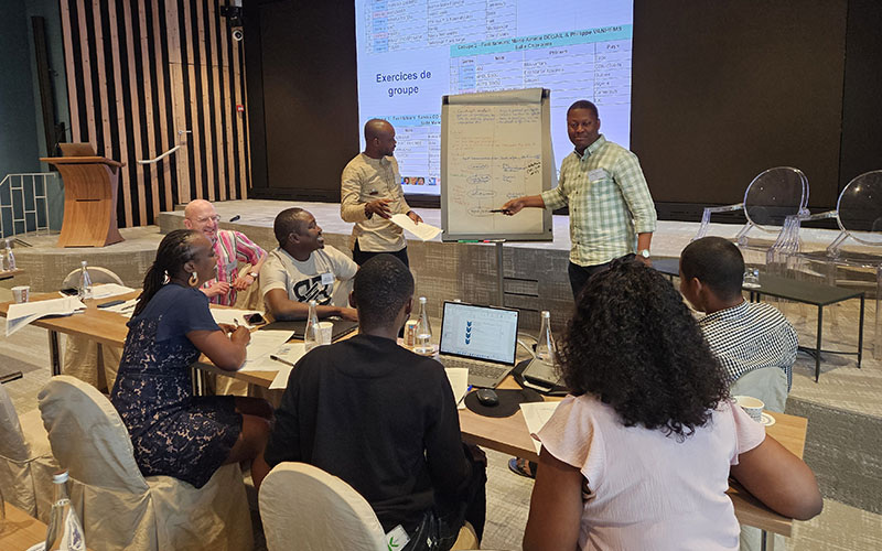 Huit personnes travaillent dans une salle de conférence. Six personnes sont assises en cercle, deux personnes se tiennent debout devant un paperboard
