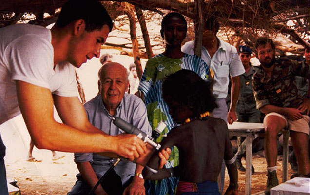 À l’extérieur, sous des arbres, Dr Charles Mérieux est assis et observe, devant lui au premier plan, un homme vacciner un enfant.