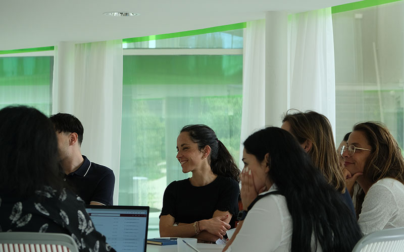 Six personnes sont assises autour d’une table et regardent vers la gauche, une femme de face apparait plus nettement que les autres