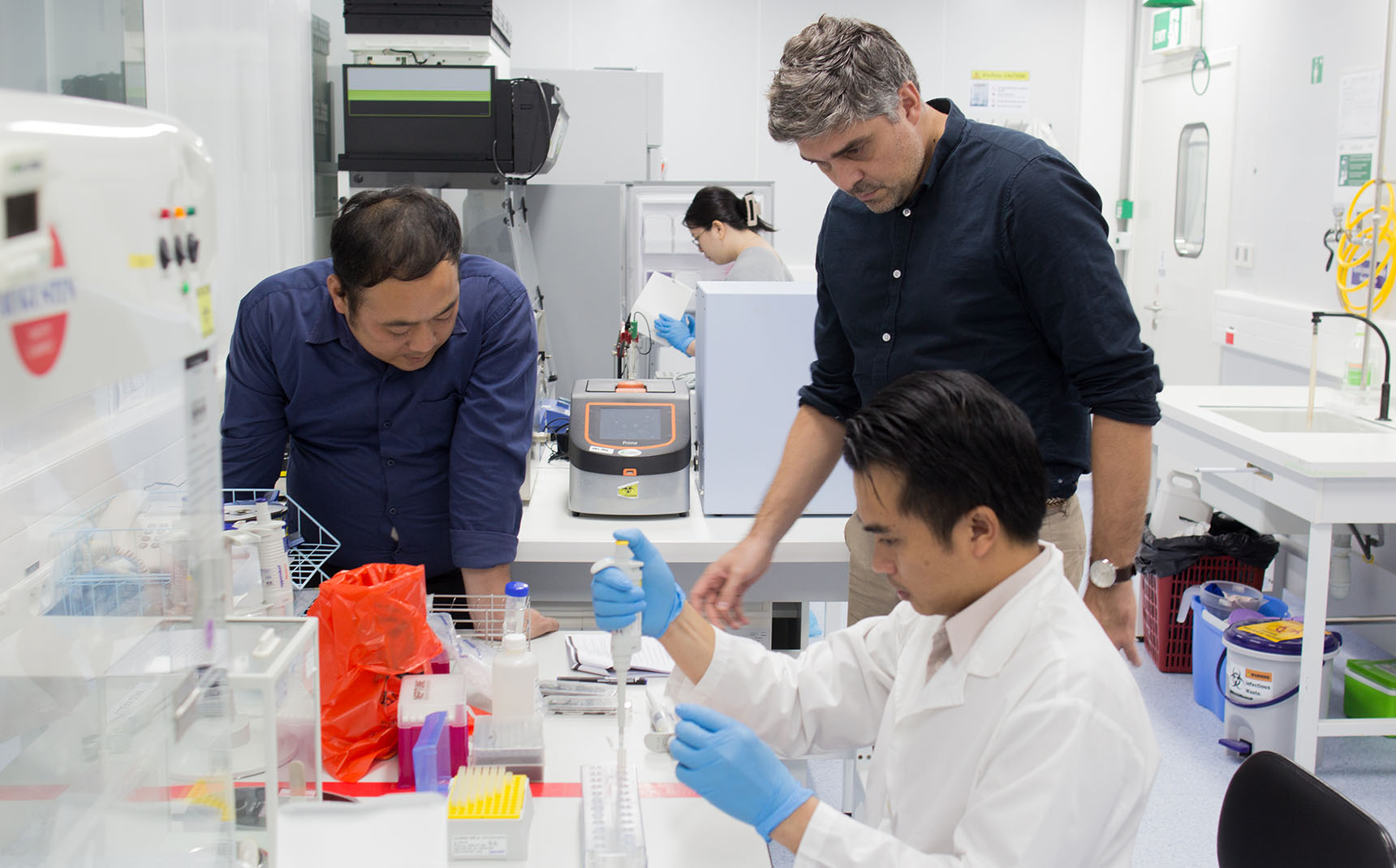 Dans un laboratoire, un homme assis manipule une pipette, au-dessus de lui se tiennent deux autres personnes, qui l’encadrent.