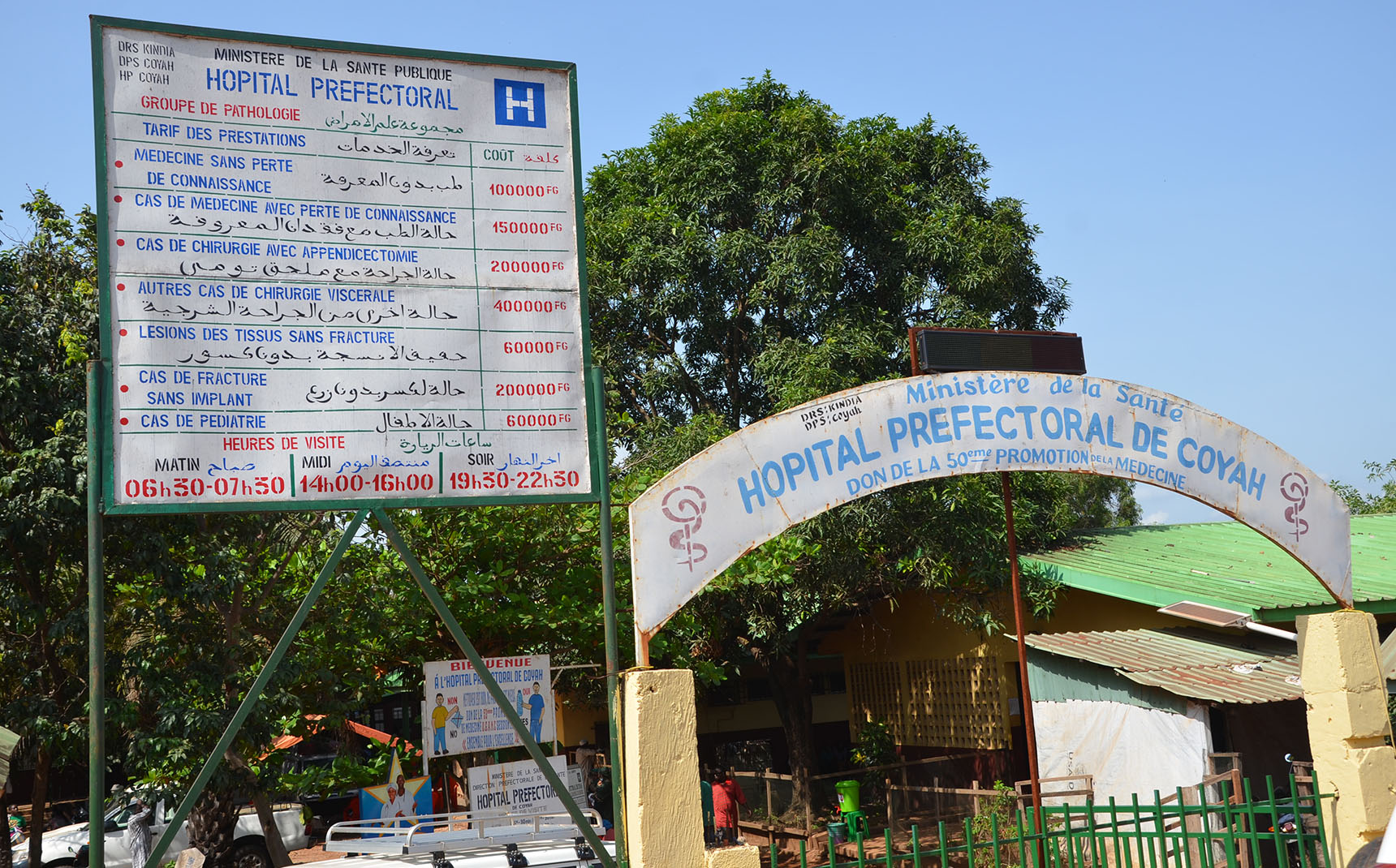 Entrée de l’hôpital, au-dessus d’un portail vert, deux pancartes indiquent le nom de l’hôpital.