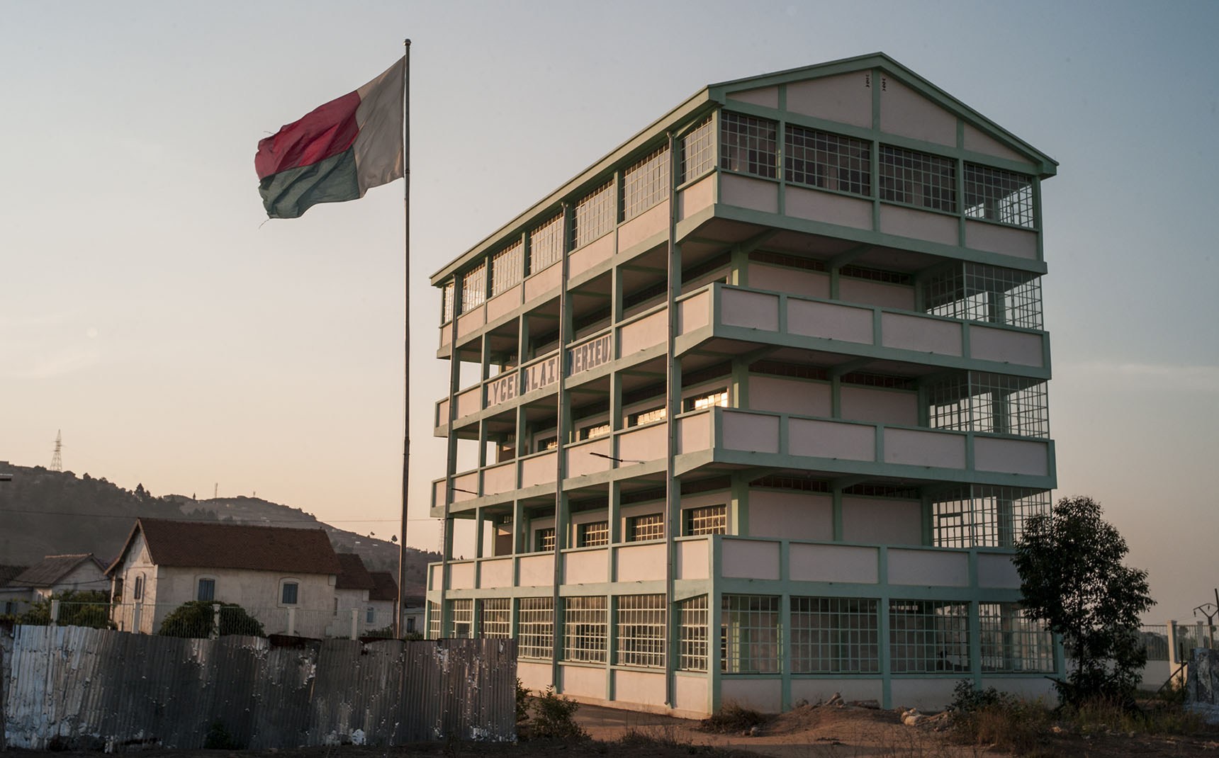 À l’extérieur, vue sur un immeuble blanc et vert sur lequel est écrit Lycée Alain Mérieux. A gauche, un drapeau de Madagascar.
