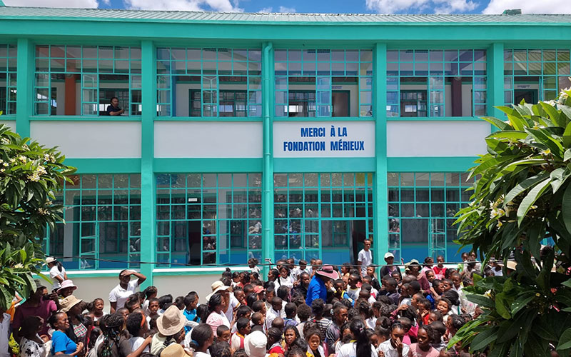 Vue de dos, une foule d'enfant se tient devant un bâtiment vert et blanc sur lequel est écrit Merci à la Fondation Mérieux.