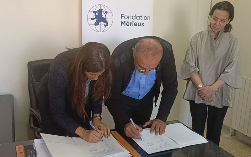 Dans un bureau, deux personnes penchées en avant pour signer des documents, en arrière plan un kakemono fait figurer le logo de la Fondation Mérieux.