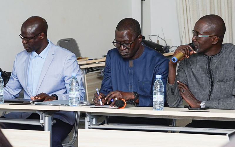 Salle de conférence avec trois participants assis de face avec une personne parlant au micro en s'adressant aux deux personnes à sa droite.
