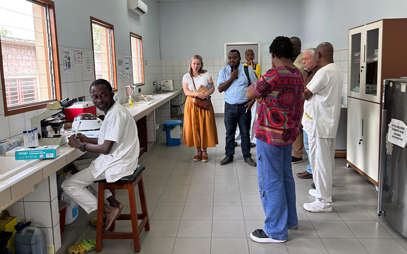 Un groupe de personnes dans un laboratoire médical, avec un technicien assis au microscope et d'autres personnes debout en discussion. Le laboratoire est bien éclairé avec des fenêtres, des équipements de laboratoire sur les plans de travail, et des armoires de rangement.