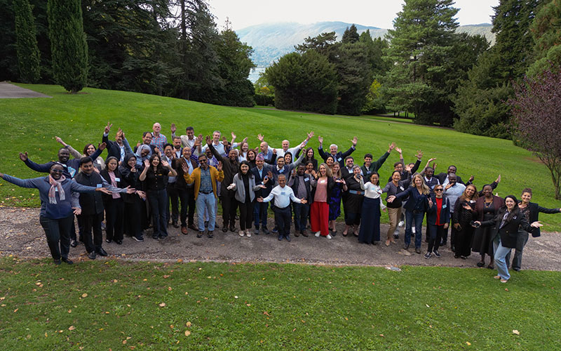 Dans un parc arboré, un groupe d’une trentaine de personnes pose, débout sur la pelouse, les bras ouverts.  